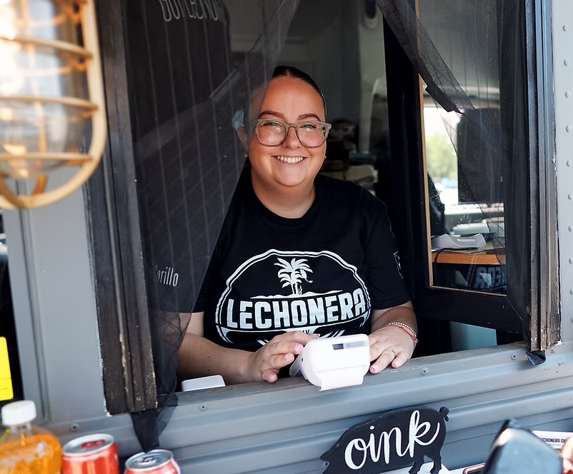 Smiling woman in a black Lechonera T‑shirt leans out of a service window, holding a card reader.