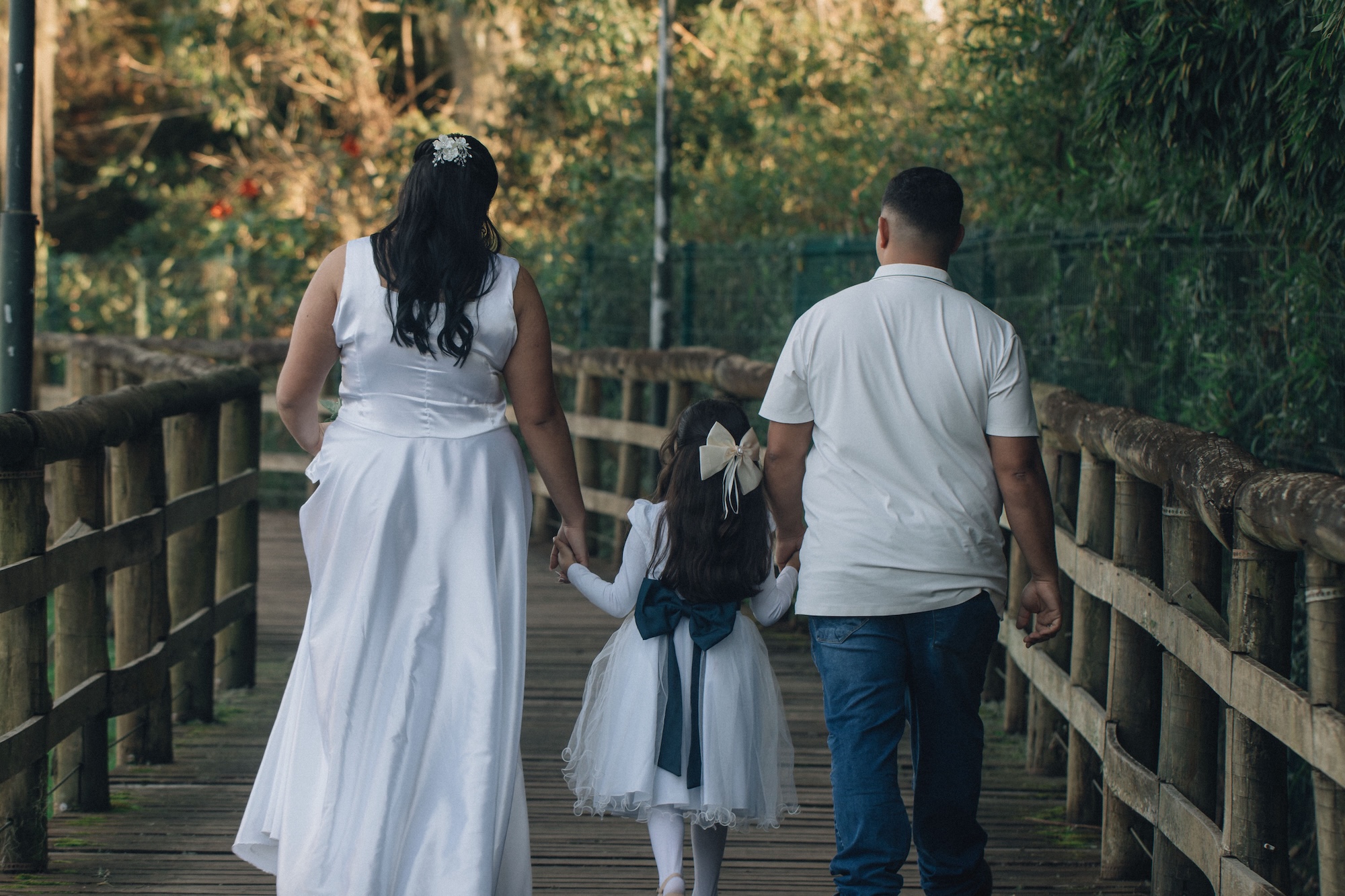 Back view of a family walking away on a wooden bridge; woman in a white dress, little girl in a white dress with a blue sash, man in a white shirt and jeans hold hands to the side.
